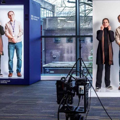 Two people posing together in a hologram tech studio, including a large printed photo of their pose in the background. Camera equipment is visible in the foreground
