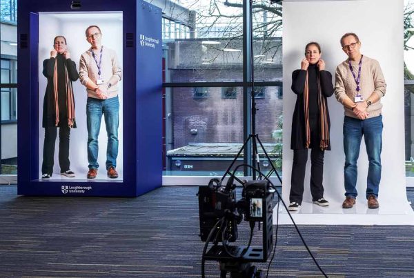 Two people posing together in a hologram tech studio, including a large printed photo of their pose in the background. Camera equipment is visible in the foreground
