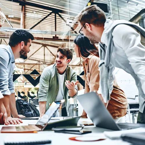 A diverse group of professionals standing around a table with laptops, engaged in a meeting or collaboration session.