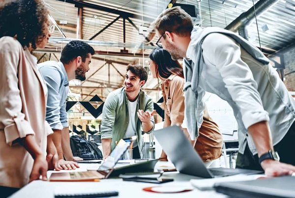 A diverse group of professionals standing around a table with laptops, engaged in a meeting or collaboration session.